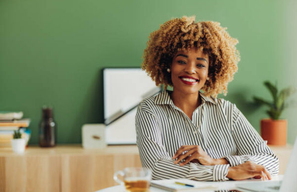 Happy African American woman looking at camera and laughing while sitting at office desk with laptop computer, notebook, pen and cup of tea.